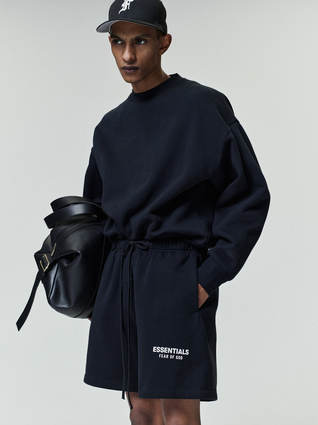 Young man wearing a black oversized sweatshirt and matching shorts with white "ESSENTIALS FEAR OF GOD" text, paired with a black baseball cap and carrying a large black leather bag.