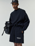 Young man wearing a black oversized sweatshirt and matching shorts with white "ESSENTIALS FEAR OF GOD" text, paired with a black baseball cap and carrying a large black leather bag.