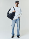 Man wearing a white long-sleeve layered shirt with "ESSENTIALS FEAR OF GOD" text on the chest, light blue straight-leg jeans, black leather shoes, a black cap, and carrying a large textured black shoulder bag.