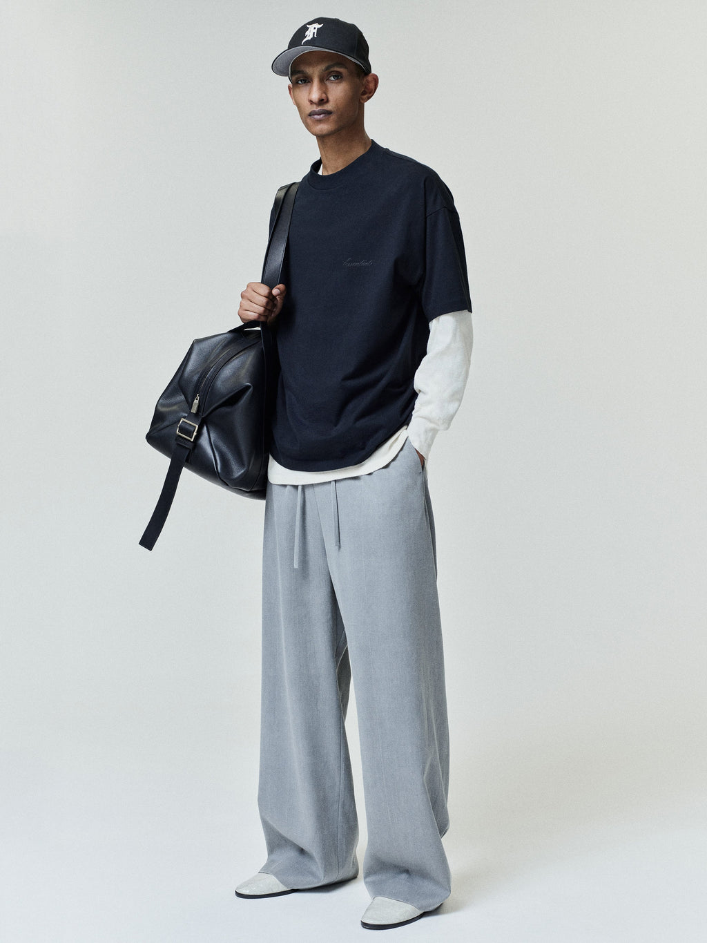 Slim man wearing a black short-sleeve t-shirt layered over a white long-sleeve shirt, loose light gray wide-leg pants, white shoes, black baseball cap, and carrying a large black leather shoulder bag.