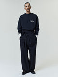 Model wearing a black long-sleeve shirt with white text on the chest layered over a white shirt, paired with high-waisted black wide-leg pants with a fabric belt and black loafers.