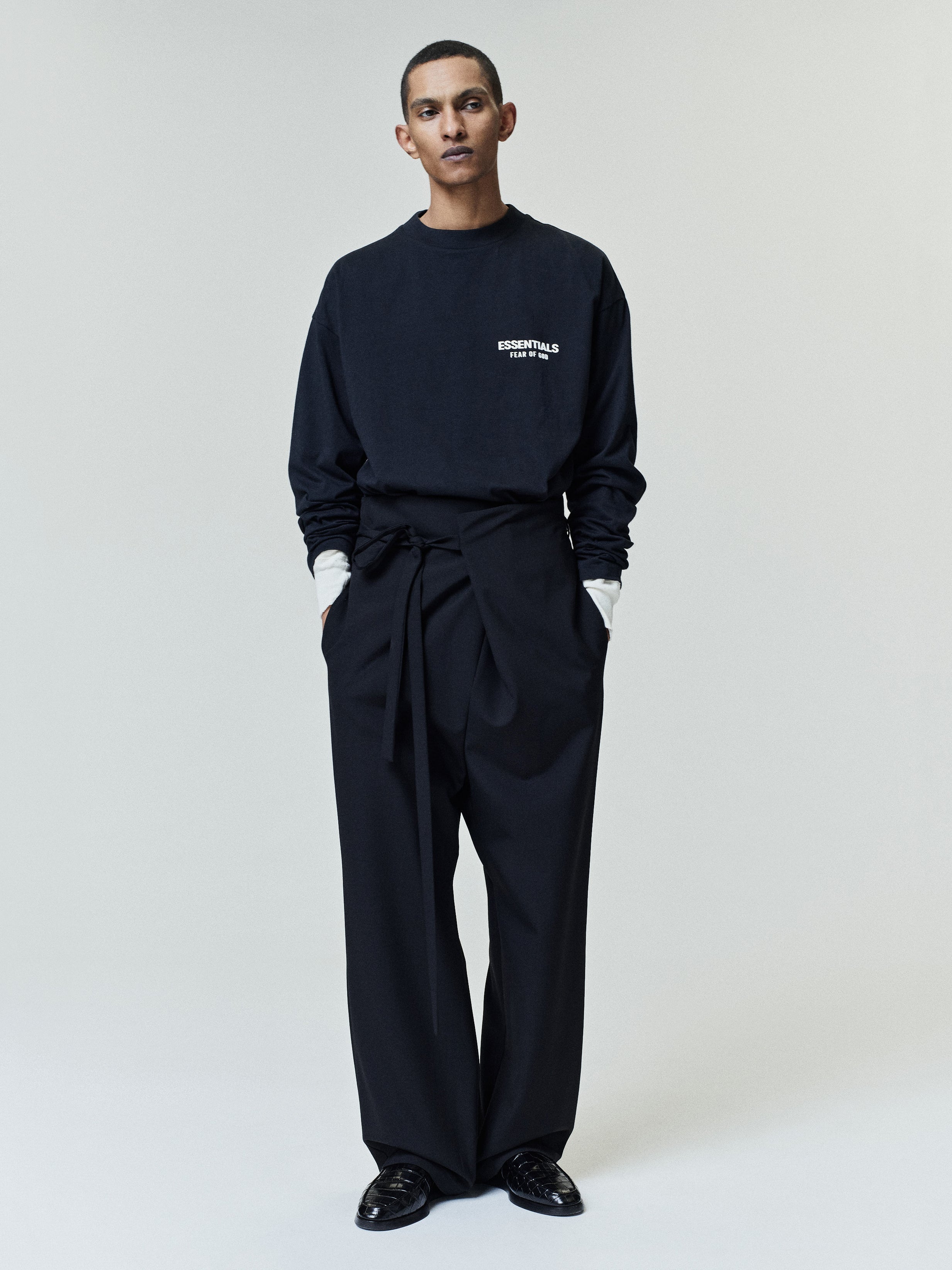 Model wearing a black long-sleeve shirt with white text on the chest layered over a white shirt, paired with high-waisted black wide-leg pants with a fabric belt and black loafers.