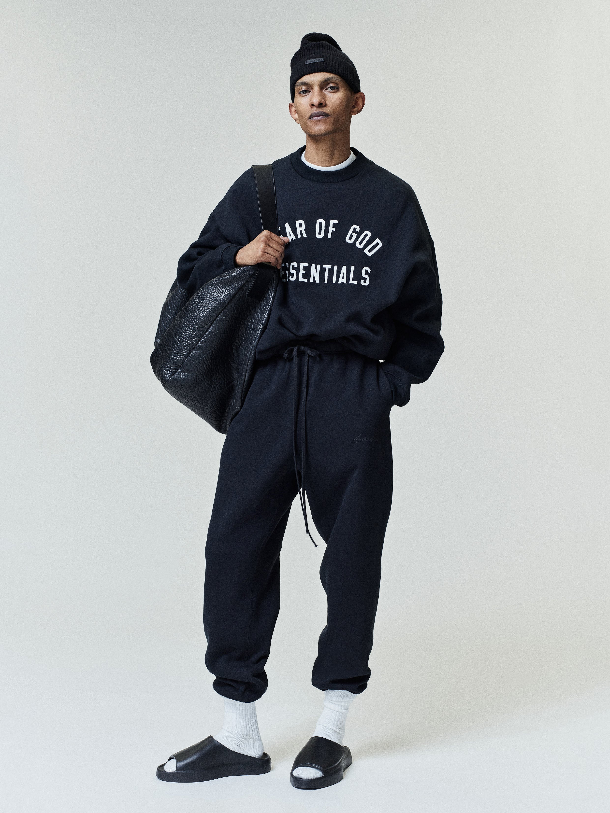 Slim man wearing a black sweatshirt with white "FEAR OF GOD ESSENTIALS" text, matching black sweatpants, white crew socks, black slides, a black beanie, and carrying a large textured black leather bag.