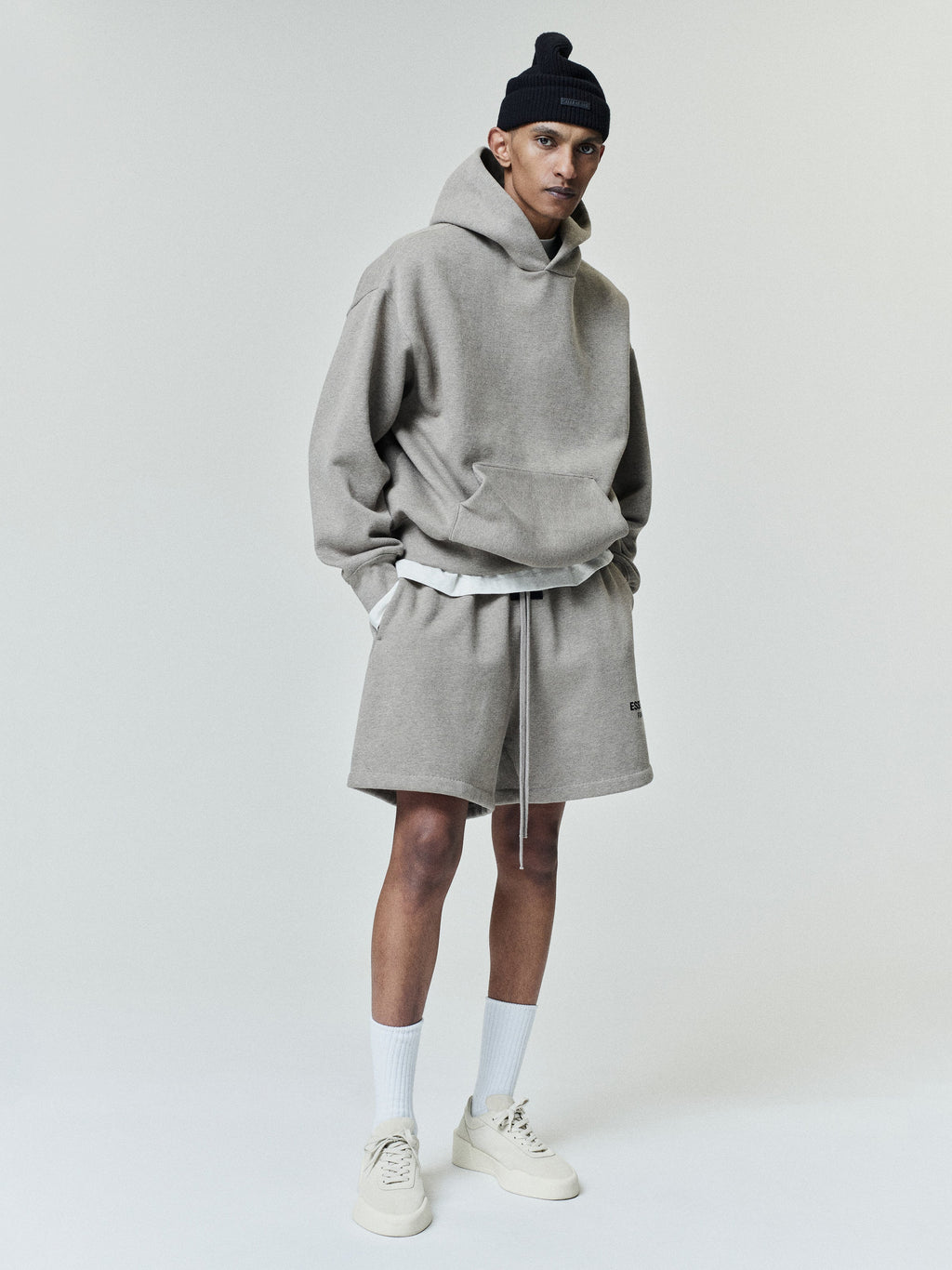 Slim man wearing a loose light gray hooded sweatshirt with long sleeves, matching drawstring shorts, white crew socks, beige sneakers, and a black knit beanie, standing with hands in pockets.