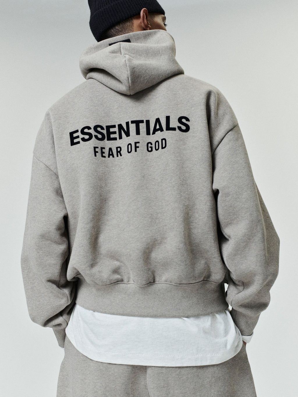 Person wearing a light gray oversized hoodie with black "ESSENTIALS FEAR OF GOD" text on the back, paired with matching pants and a black knit beanie.