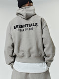 Person wearing a light gray oversized hoodie with black "ESSENTIALS FEAR OF GOD" text on the back, paired with matching pants and a black knit beanie.