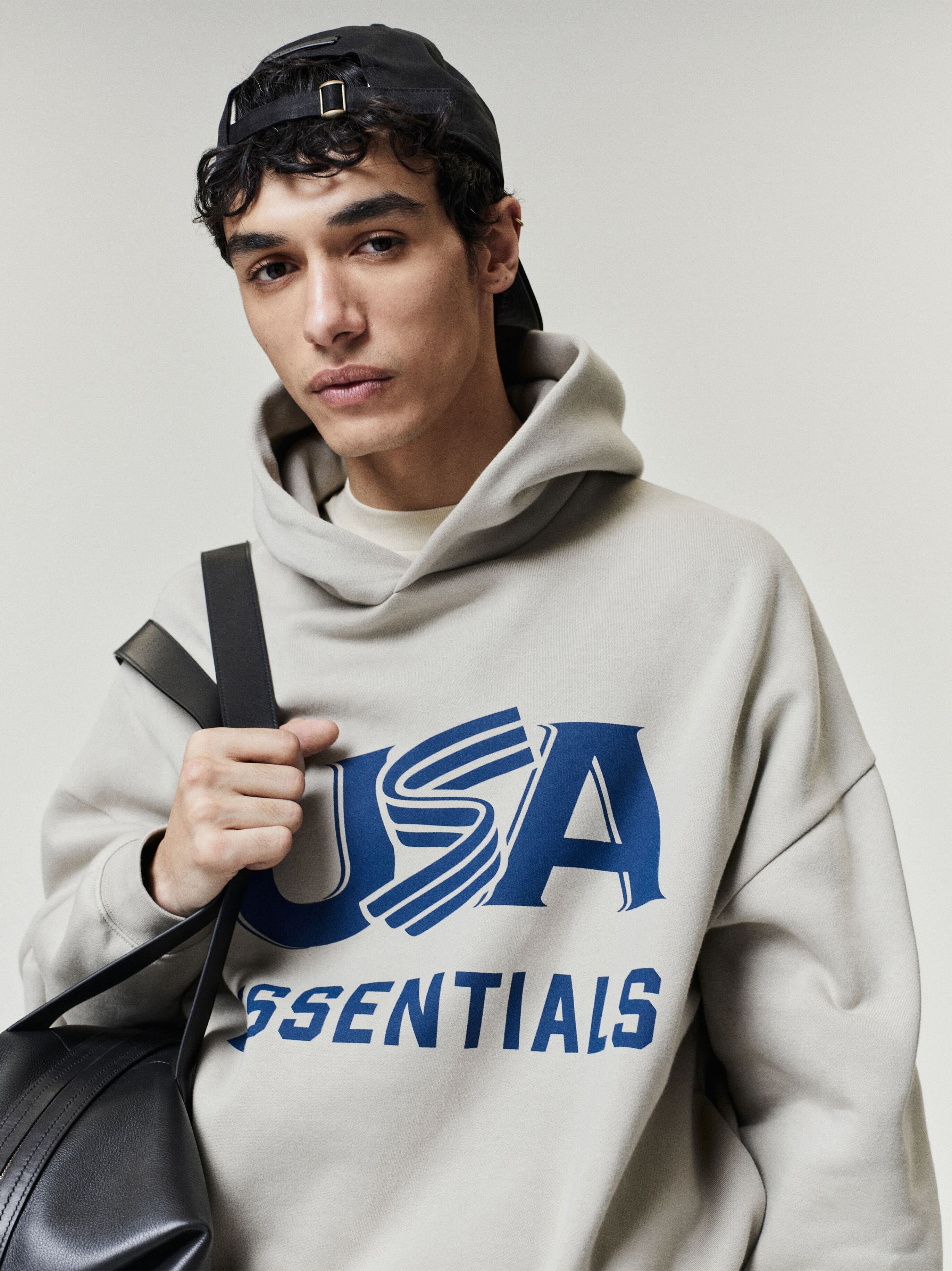 Young man wearing light gray hoodie with blue "USA ESSENTIALS" print, black cap worn backward, and carrying black backpack strap