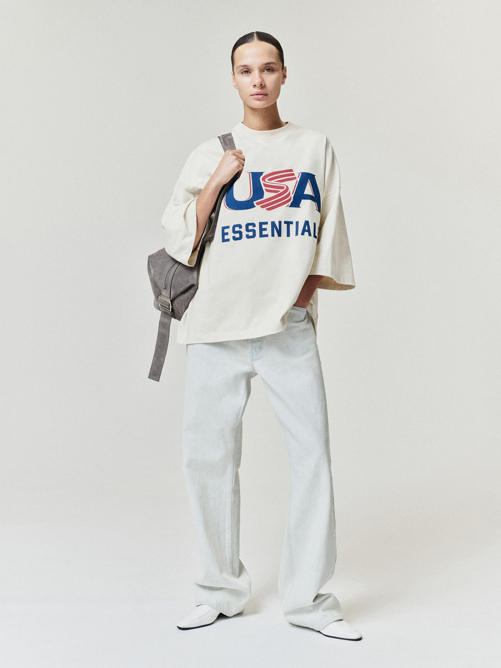 Model wearing oversized off-white t-shirt with red and blue USA print, light-colored wide-leg pants, and carrying gray backpack