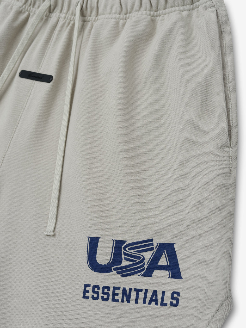 Close-up of light beige sweatpants with elastic waistband, drawstrings, side pocket, and navy "USA ESSENTIALS" print on leg