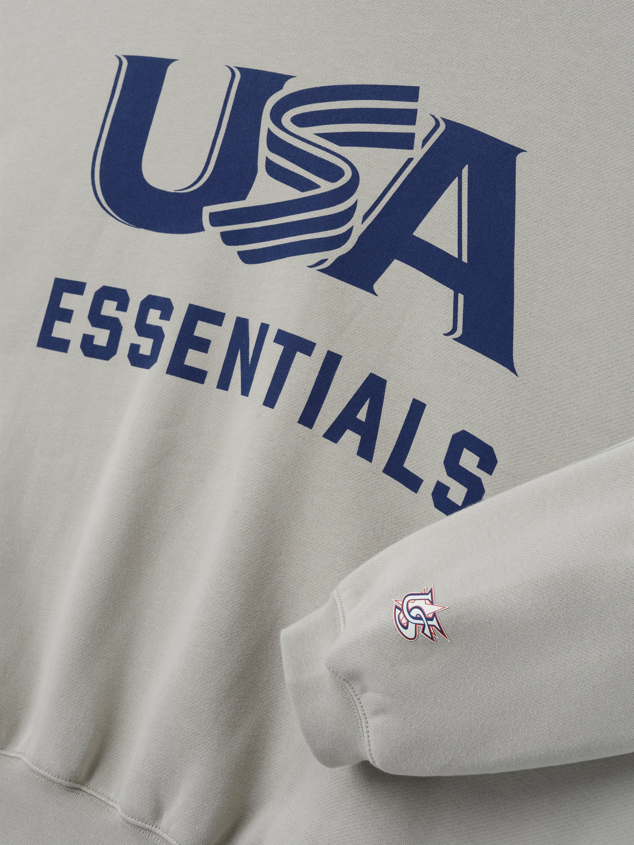 Close-up of cream-colored sweatshirt with navy "USA" graphic and "ESSENTIALS" text and small logo on sleeve cuff edge