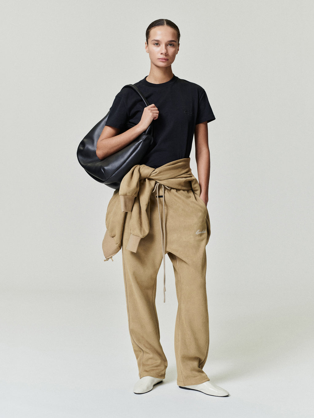 Person wearing a black t-shirt and beige pants holding a black bag on a light gray background