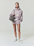 female model wearing lavender cropped raglan hoodie with ESSENTIALS Fear of God text and matching shorts, carrying grey bag