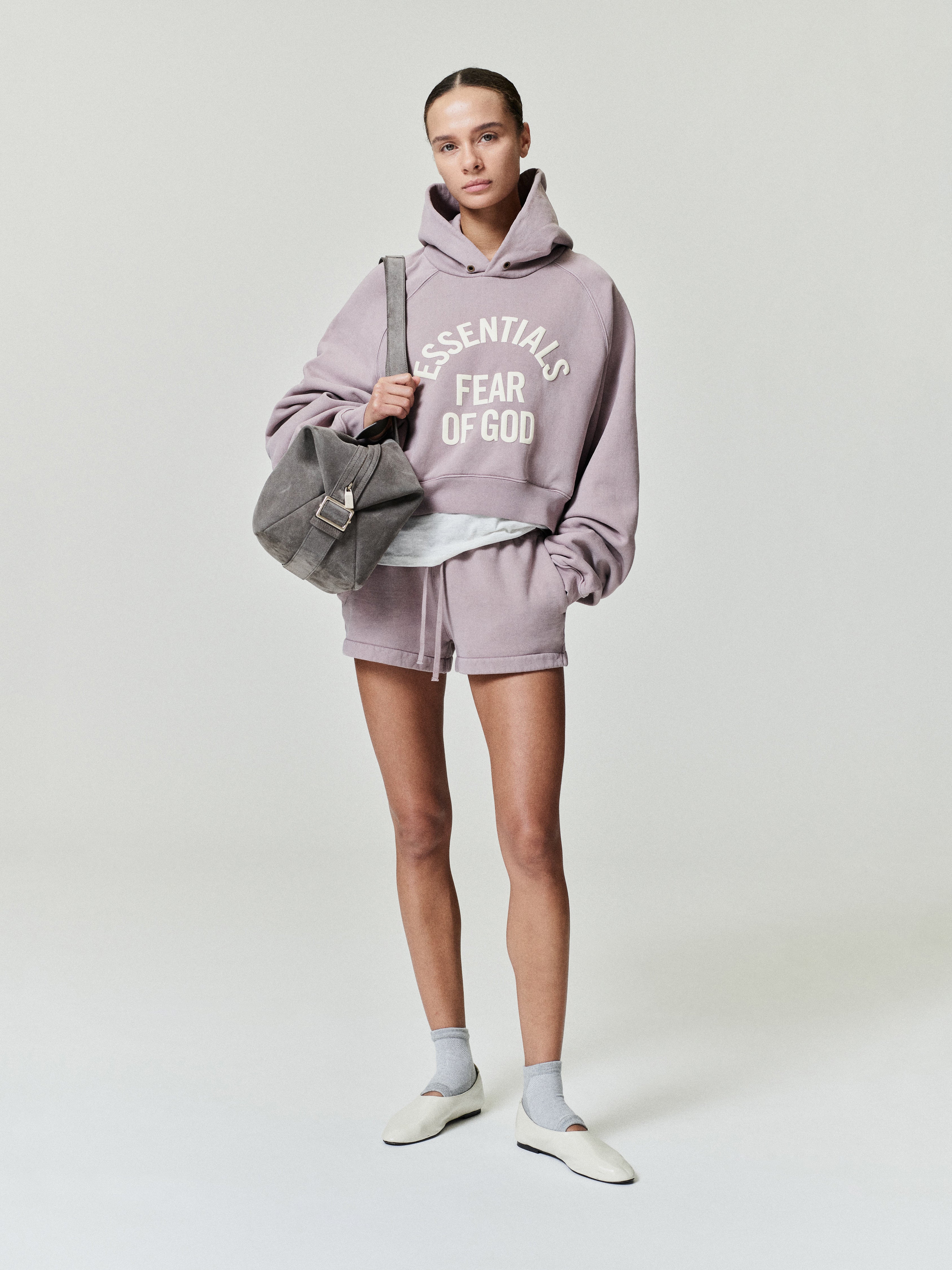 female model wearing lavender cropped raglan hoodie with ESSENTIALS Fear of God text and matching shorts, carrying grey bag