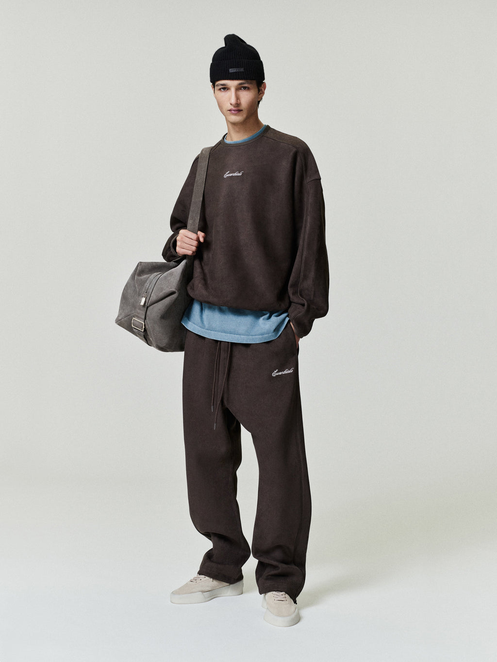 Person wearing a brown sweatshirt and pants with a gray bag on a white background