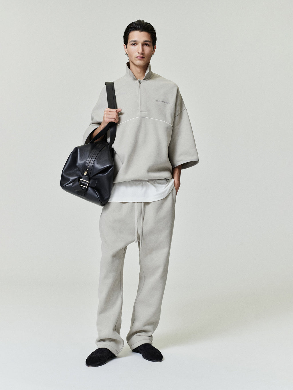 man wearing light gray cropped fleece polo sweatshirt with wide 3/4 sleeves and matching relaxed fleece pants carrying black leather bag
