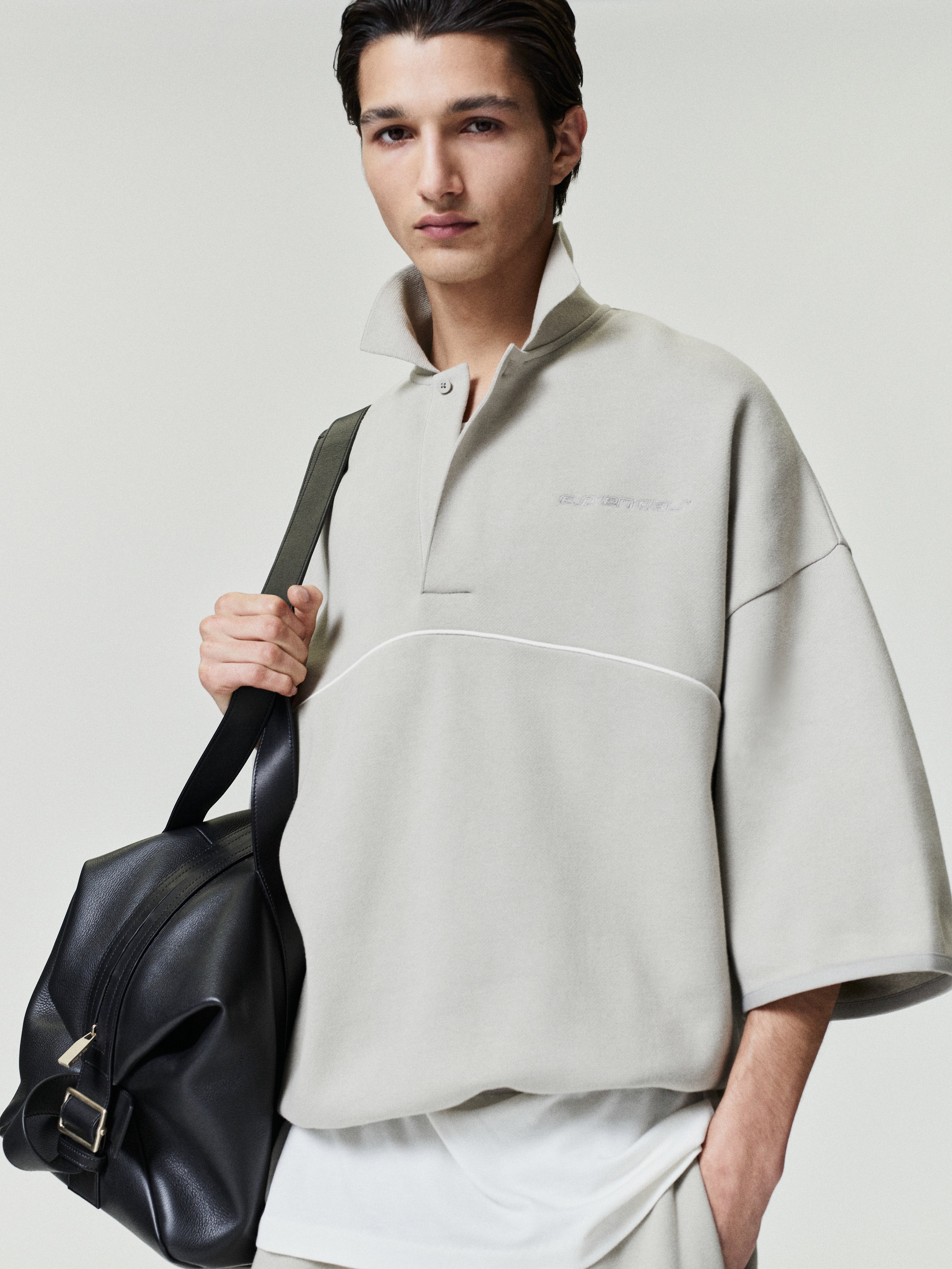 man wearing light beige cropped polo sweatshirt with 3/4 sleeves and subtle embroidered logo holding black leather bag